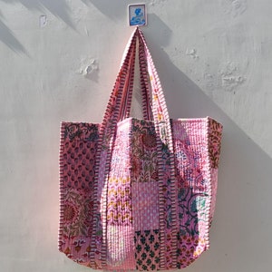 May include: A pink patchwork tote bag with floral patterns and white stripes. The bag has a quilted texture and is made of cotton fabric.