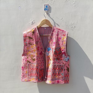 Op de afbeelding: Een roze gewatteerd vest met een bloemenprint en een gestreepte voering. Het vest heeft twee zakken en een knoopsluiting.