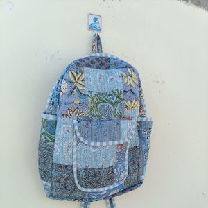 Op de afbeelding: Een blauwe en witte patchwork-rugzak van gestikt materiaal met een bloemenpatroon. De rugzak heeft een groot vak aan de voorkant en twee verstelbare banden.