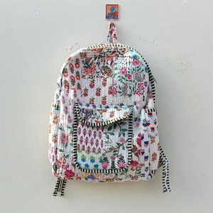 以下が含まれることがあります： ピンク、赤、青の花が咲く、カラフルな花柄の白いキルティングバックパック。 バックパックにはフロントポケットと緑のストライプのトリムが付いています。 白い壁に掛けられています。