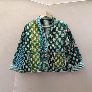 May include: A quilted jacket featuring a patchwork design in shades of dark green, olive, and teal. The garment has a V-neckline, long sleeves with folded cuffs, two front patch pockets, and a button-front closure with a striped interior lining.