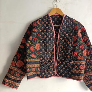 Peut inclure: Veste matelassée bleu marine ornée de motifs floraux rouges et oranges. Bordée de rose, elle possède deux poches. Suspendue à un cintre en bois, cette veste est un vêtement élégant.