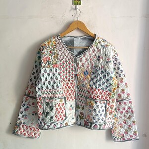 Puede incluir: Chaqueta acolchada con un diseño de patchwork con estampados florales en varios colores. La chaqueta tiene cuello en V, dos bolsillos y un forro a rayas. Los bordes están ribeteados con una tela a rayas en blanco y negro.