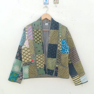 Puede incluir: Una chaqueta patchwork en tonos verdes, azules y rosas, colgada de una percha de madera. La chaqueta presenta una mezcla de telas estampadas, incluyendo diseños florales y geométricos. El frente abierto revela un forro a rayas.