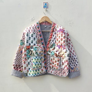 以下が含まれることがあります： ピンク、ブルー、グリーン、ブラウンの色合いの花柄と幾何学模様がパッチワークされた、白いキルティングジャケット。Vネック、長袖、ボタン留めです。