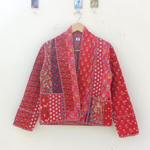 Può includere: Una giacca trapuntata rossa con stampa floreale e un lungo davanti aperto. La giacca ha un design patchwork con diversi motivi floreali in tonalità di rosso, rosa, blu e giallo.