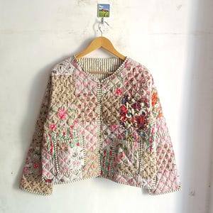 May include: A quilted jacket with a patchwork design. The jacket features a V-neck, two pockets, and a variety of floral patterns in shades of pink, red, and beige. The jacket is hanging on a wooden hanger.