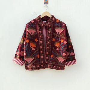 May include: A deep burgundy jacket featuring intricate floral embroidery in shades of pink, orange, and green. The garment has long sleeves with folded cuffs revealing a white and red striped lining, a high collar, and a matching striped interior.