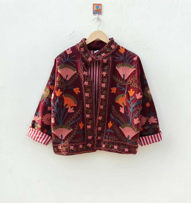 May include: A deep burgundy jacket featuring intricate floral embroidery in shades of pink, orange, and green. The garment has long sleeves with folded cuffs revealing a white and red striped lining, a high collar, and a matching striped interior.