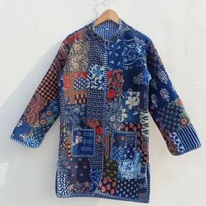 Può includere: Una lunga giacca patchwork con collo alla coreana. La giacca presenta una varietà di toppe in tessuto blu, rosso e crema con motivi floreali e geometrici. La giacca è appesa a una gruccia di legno.