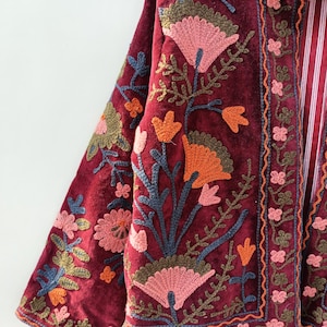 May include: A deep burgundy velvet jacket featuring intricate floral embroidery in shades of pink, orange, and olive green. The garment has long sleeves, a high collar, and a vertical front placket with a red and white striped lining visible inside.
