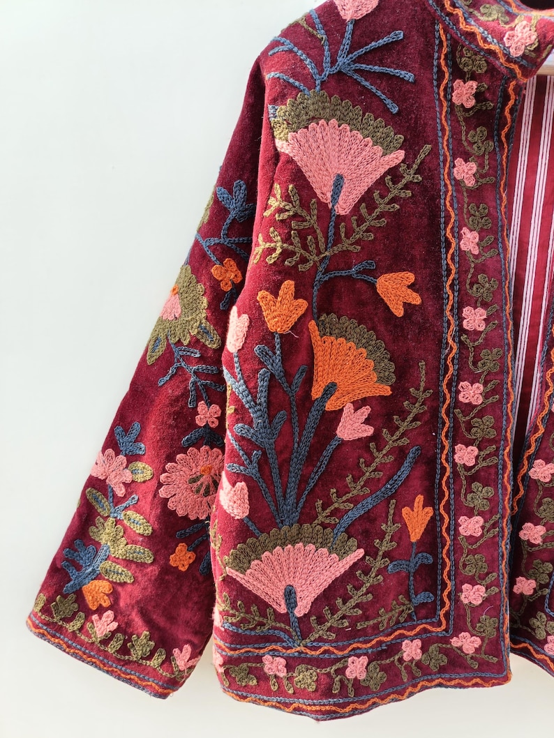 May include: A deep burgundy velvet jacket featuring intricate floral embroidery in shades of pink, orange, and olive green. The garment has long sleeves, a high collar, and a vertical front placket with a red and white striped lining visible inside.