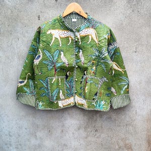 May include: Green quilted jacket with a jungle-themed print featuring leopards, birds, and palm trees. The jacket has a mandarin collar, button closure, and striped trim. The jacket is hanging on a wooden hanger.