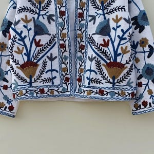 May include: White embroidered jacket with a floral pattern in shades of blue, brown, and red. The jacket has a long sleeve and a button-up front.
