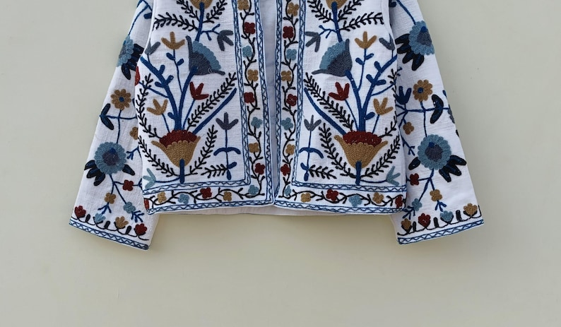 May include: White embroidered jacket with a floral pattern in shades of blue, brown, and red. The jacket has a long sleeve and a button-up front.