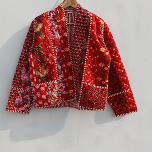 May include: A red quilted jacket with floral and geometric patterns. The jacket has a patchwork design with various floral prints and a striped interior lining. It is hanging on a wooden hanger against a white background.