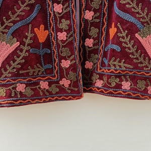 May include: A deep burgundy velvet jacket featuring intricate floral embroidery in shades of pink, orange, blue, and olive green. The design includes large stylised flower motifs and delicate vine patterns along the hem and front panels.