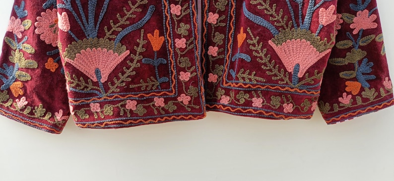 May include: A deep burgundy velvet jacket featuring intricate floral embroidery in shades of pink, orange, blue, and olive green. The design includes large stylised flower motifs and delicate vine patterns along the hem and front panels.