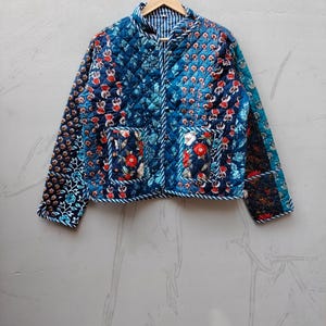 May include: A quilted jacket featuring a patchwork design with various blue, red, and white floral patterns. The jacket has long sleeves, a front button closure, two front pockets, and a blue and white striped trim along the edges and collar.