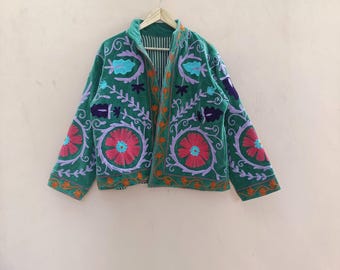 Puede incluir: Una chaqueta verde con bordados florales coloridos. La chaqueta tiene un cuello alto y está adornada con diseños florales rojos, rosas, morados y turquesas. La chaqueta está colgada de una percha de madera sobre un fondo melocotón claro.