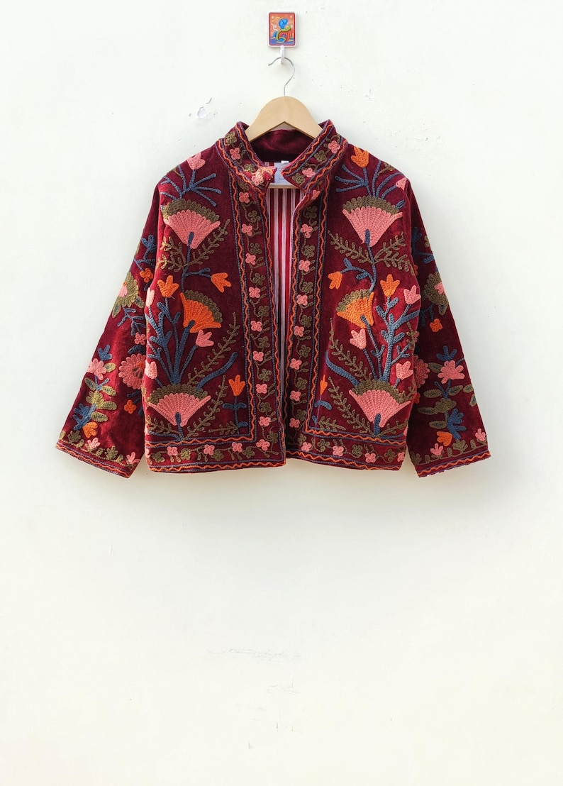 May include: A deep burgundy velvet jacket featuring intricate floral embroidery in shades of pink, orange, and blue. The jacket has a high collar, long sleeves, and a front opening that reveals a red and white striped lining on the interior.