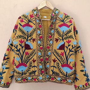 May include: Mustard yellow jacket with detailed floral embroidery in blue, pink, and burgundy. The jacket has a stand-up collar and is displayed on a wooden hanger. The embroidery design is symmetrical and covers the front and sleeves.