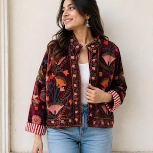 May include: A deep burgundy velvet jacket featuring intricate floral embroidery in shades of orange, pink, and green. The jacket has a mandarin collar, long sleeves with red and white striped cuffs, and a front opening worn over a white top.