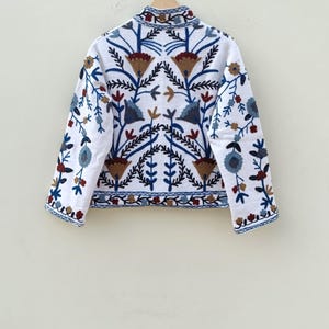 May include: White embroidered jacket with a floral pattern in shades of blue, brown, and red. The jacket has a stand-up collar and long sleeves.