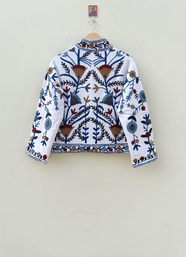May include: White embroidered jacket with a floral pattern in shades of blue, brown, and red. The jacket has a stand-up collar and long sleeves.