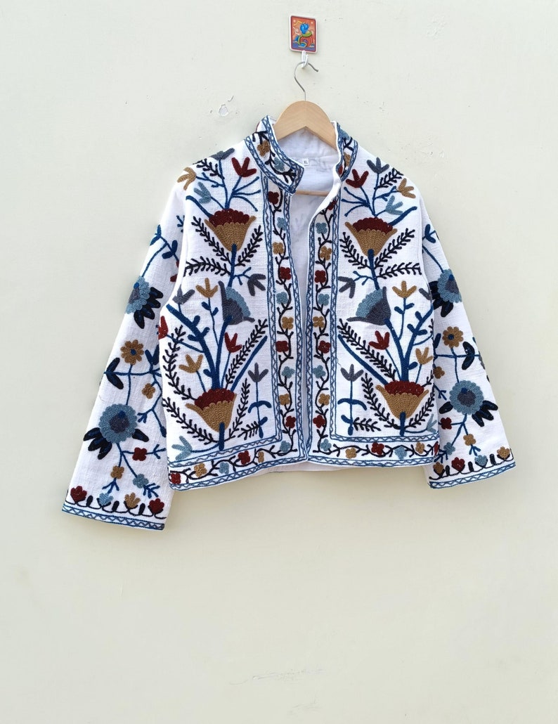May include: White embroidered jacket with a floral pattern in shades of blue, brown, and red. The jacket has a mandarin collar and a button closure.