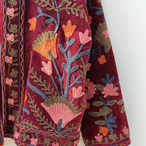 May include: A deep burgundy velvet jacket featuring intricate floral embroidery in shades of pink, orange, blue, and olive green. The garment displays detailed stitching along the front placket and sleeves with a dense botanical pattern.