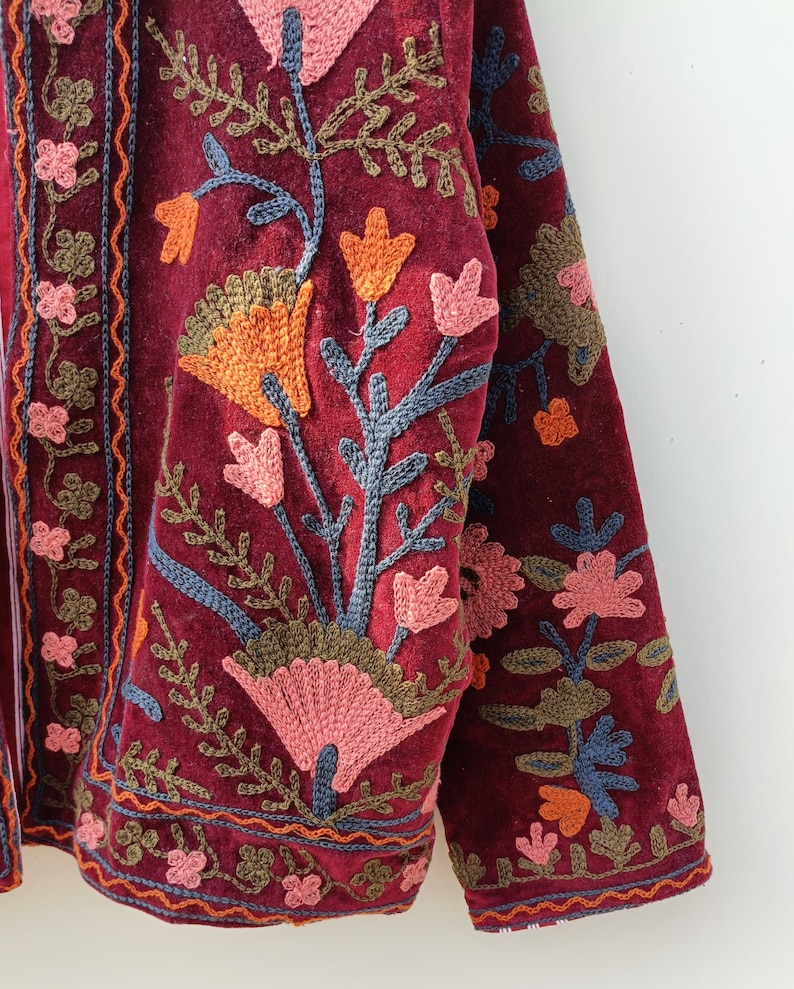 May include: A deep burgundy velvet jacket featuring intricate floral embroidery in shades of pink, orange, blue, and olive green. The garment displays detailed stitching along the front placket and sleeves with a dense botanical pattern.