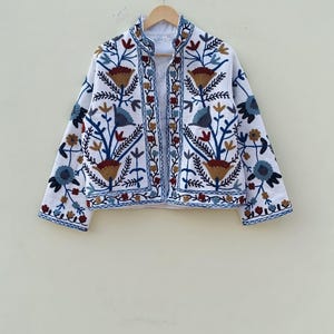 May include: White embroidered jacket with a floral pattern in shades of blue, brown, and red. The jacket has a mandarin collar and long sleeves.