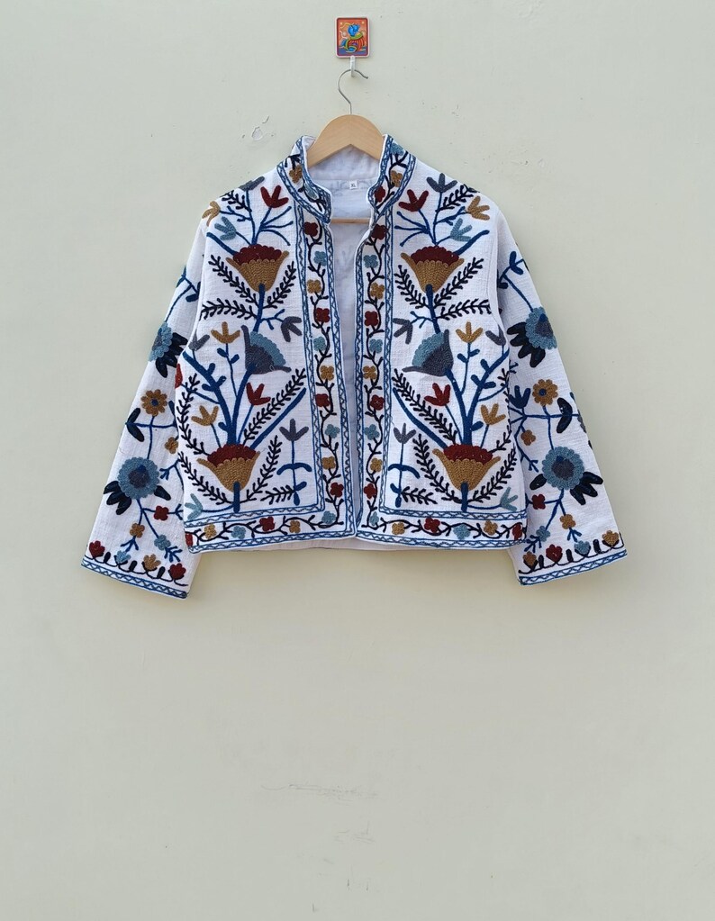 May include: White embroidered jacket with a floral pattern in shades of blue, brown, and red. The jacket has a mandarin collar and long sleeves.
