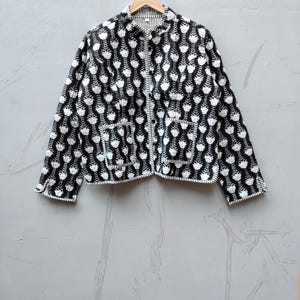 May include: A long-sleeved, button-down jacket featuring a repeating black and white floral pattern. The garment includes a mandarin collar, two front patch pockets, and visible white stitching along the edges of the cuffs, hem, and pockets.