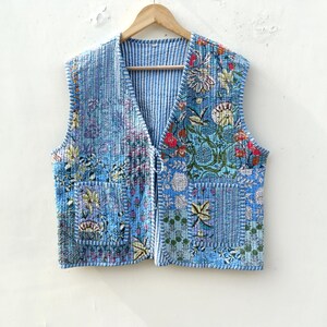 May include: A blue quilted vest with a floral pattern and two front pockets. The vest has a V-neck and is hanging on a wooden hanger. The floral pattern includes red, yellow, and green flowers. The vest is approximately 61 cm long.