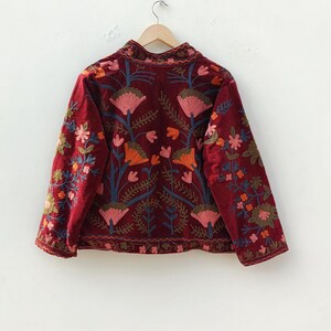 May include: A deep burgundy velvet jacket featuring intricate floral embroidery in shades of pink, orange, and blue. The jacket has long sleeves, a high collar, and a button-down front, with a repeating botanical pattern across the entire garment.