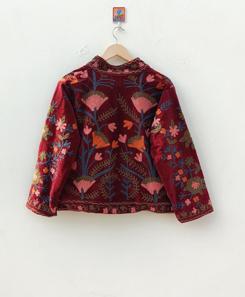 May include: A deep burgundy velvet jacket featuring intricate floral embroidery in shades of pink, orange, and blue. The jacket has long sleeves, a high collar, and a button-down front, with a repeating botanical pattern across the entire garment.