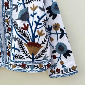 May include: A white linen jacket with colorful floral embroidery. The embroidery features blue, red, brown, and gold thread. The jacket has a long sleeve and a button closure.