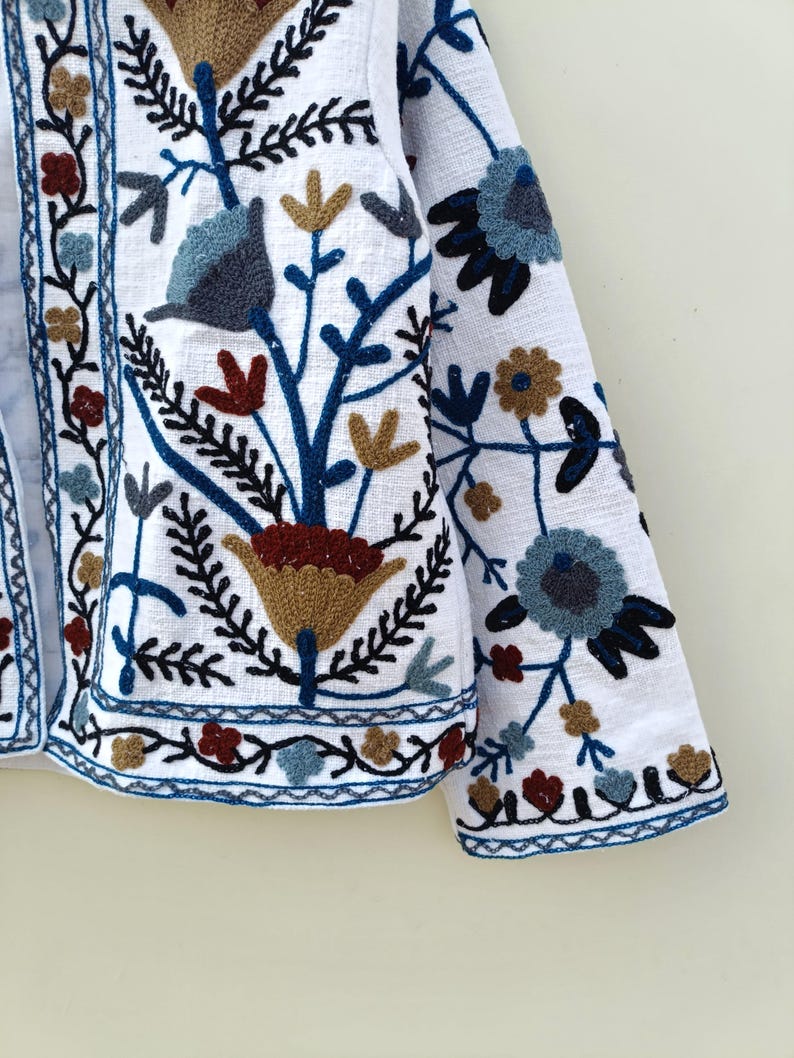 May include: A white linen jacket with colorful floral embroidery. The embroidery features blue, red, brown, and gold thread. The jacket has a long sleeve and a button closure.