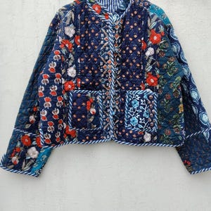 Peut inclure: Une veste matelassée sur fond bleu foncé avec un motif floral multicolore en rouge, blanc et bleu clair. Le vêtement présente une encolure ronde, des manches longues, deux poches plaquées à l'avant et une patte centrale avec un liseré rayé bleu et blanc.