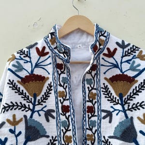 May include: White cotton jacket with embroidered floral designs in shades of blue, brown, and gold. The jacket has a mandarin collar and a front closure.