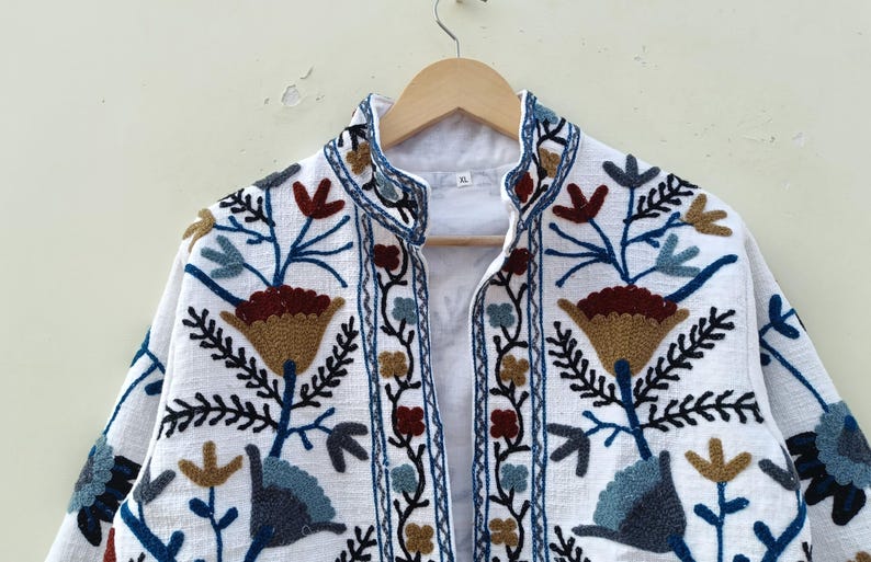 May include: White cotton jacket with embroidered floral designs in shades of blue, brown, and gold. The jacket has a mandarin collar and a front closure.