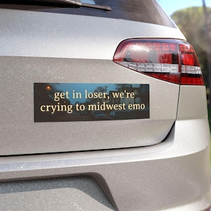 May include: A rectangular bumper sticker with the text "get in loser, we're crying to midwest emo" in a gold font. The background of the sticker is a blurry image of a street scene. The sticker is affixed to the back of a silver car.