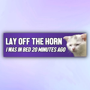 May include: A purple and white bumper sticker with a white cat with a sad expression. The text on the sticker reads "Lay off the horn I was in bed 20 minutes ago."