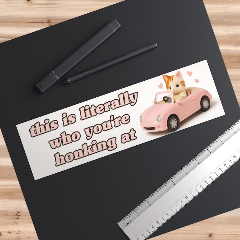 May include: A white sticker with the text "this is literally who you're honking at" in pink letters. The sticker features a pink toy car with a cat inside. A black marker and pen are on the top left.
