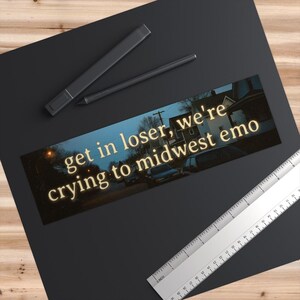 May include: A rectangular sticker with the text "get in loser, we're crying to midwest emo" in a glowing yellow font. The background shows a street scene with houses and cars.