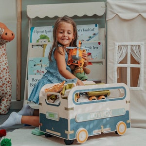 Puede incluir: Una ni&ntilde;a juega con un helic&oacute;ptero de juguete y otros juguetes en una caja de juguetes azul y blanca con la palabra "Toybox" impresa en el lateral. La caja de juguetes tiene ruedas y est&aacute; llena de juguetes.
