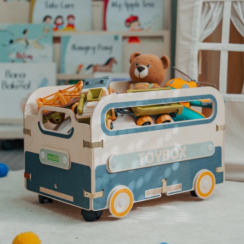 Puede incluir: Una caja de juguetes azul y blanca con forma de autob&uacute;s vintage con ruedas. La caja de juguetes est&aacute; llena de varios juguetes, incluyendo un oso de peluche, un cami&oacute;n de juguete y un coche de juguete. La caja de juguetes tiene la palabra "TOYBOX" impresa en el lateral.