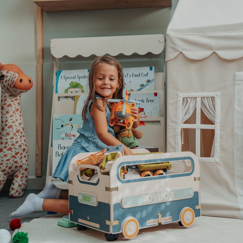 Puede incluir: Una ni&ntilde;a juega con un helic&oacute;ptero de juguete y otros juguetes en una caja de juguetes azul y blanca con la palabra "Toybox" impresa en el lateral. La caja de juguetes tiene ruedas y est&aacute; llena de juguetes.